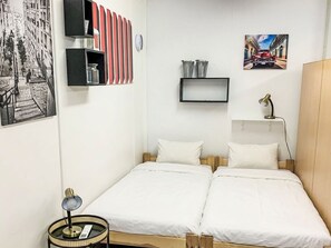 A room with two beds, a small table with a lamp, a shelf with decorative items, and a framed picture on the wall.