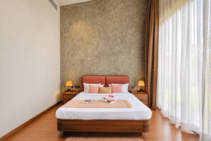 Une chambre d’hôtel avec un grand lit, une tête de lit en bois, deux tables de chevet avec des lampes, une table d’appoint et une fenêtre avec des rideaux.