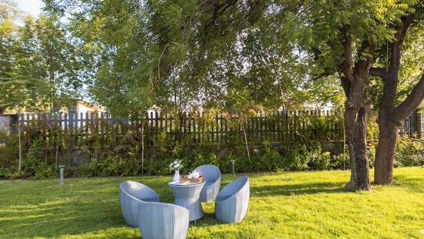 Un espacio al aire libre con sillas de mimbre y una mesa, rodeado de abundante vegetación y una valla de madera.