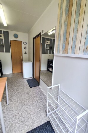 A compact room with a door, a small white refrigerator, a black mat, a white shelf, and a mirror with a wooden frame.
