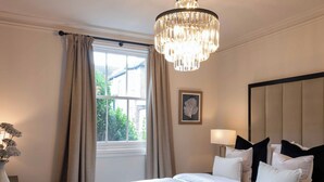 Habitación de hotel con una cama grande, un candelabro, una ventana con cortinas y una mesita de noche con lámpara.