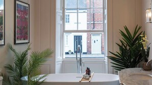 Un baño con una bañera independiente, una ventana con vista a un edificio y una planta en maceta.