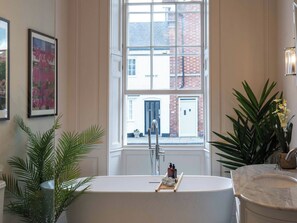 Ein Badezimmer mit freistehender Badewanne, einem Fenster mit Blick auf ein Gebäude und einer Zimmerpflanze.