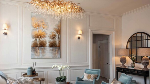 Un salón bien iluminado con una araña, un cuadro abstracto de gran tamaño, dos sillones, una mesa de centro y una mesita auxiliar con un jarrón de flores.