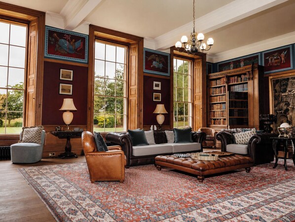 Een ruime kamer met een groot raam, een leren zithoek, een kamerbreed tapijt met dessin en een boekenplank vol boeken.