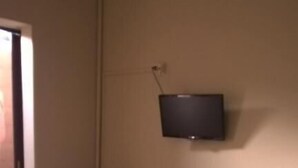 Quarto com uma cama, uma mesinha, televisão fixada na parede e uma porta que dá para outra área.
