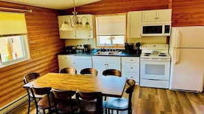 A compact kitchen with wooden walls, a dining table with chairs, a sink, a stove, and a refrigerator. - 1818 Trailside Cedar - Cabin 2 (Grand Marais)