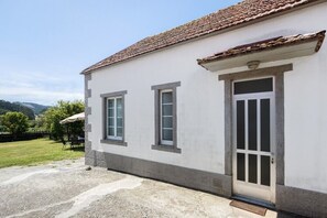 Ein zweistöckiges Gebäude mit weißer Fassade und einem Ziegeldach. Im Erdgeschoss befinden sich zwei Fenster und eine Tür mit Fenster. Das Gebäude wird von Grünflächen und Hügeln im Hintergrund begleitet.