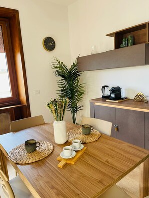 Un'area pranzo con un tavolo in legno, sedie, un vaso con fiori, una macchina da caffè e un orologio appeso al muro.