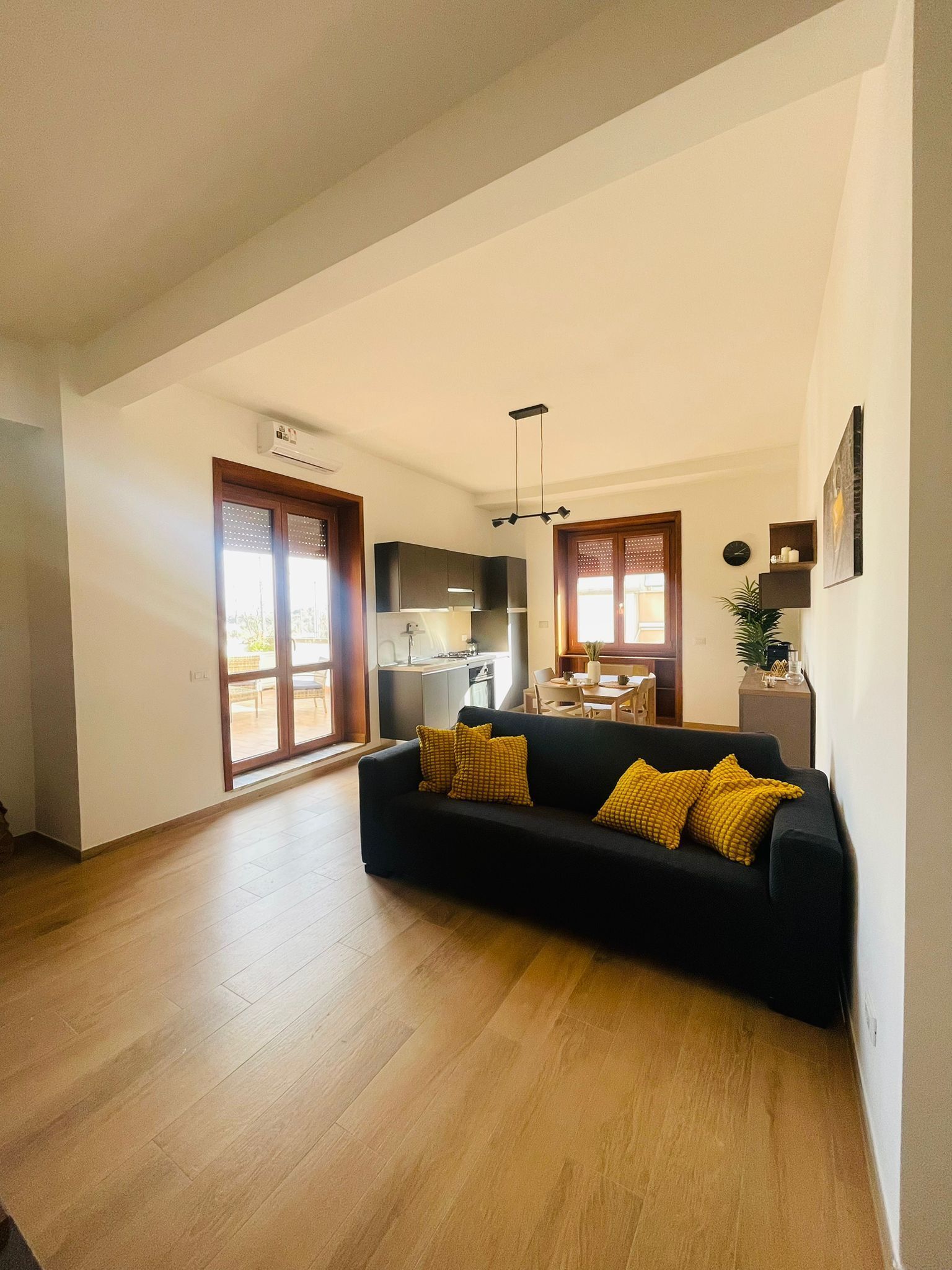 A spacious living area with a black sofa and wooden flooring, a kitchen in the background, and a sliding glass door leading to an outdoor area.