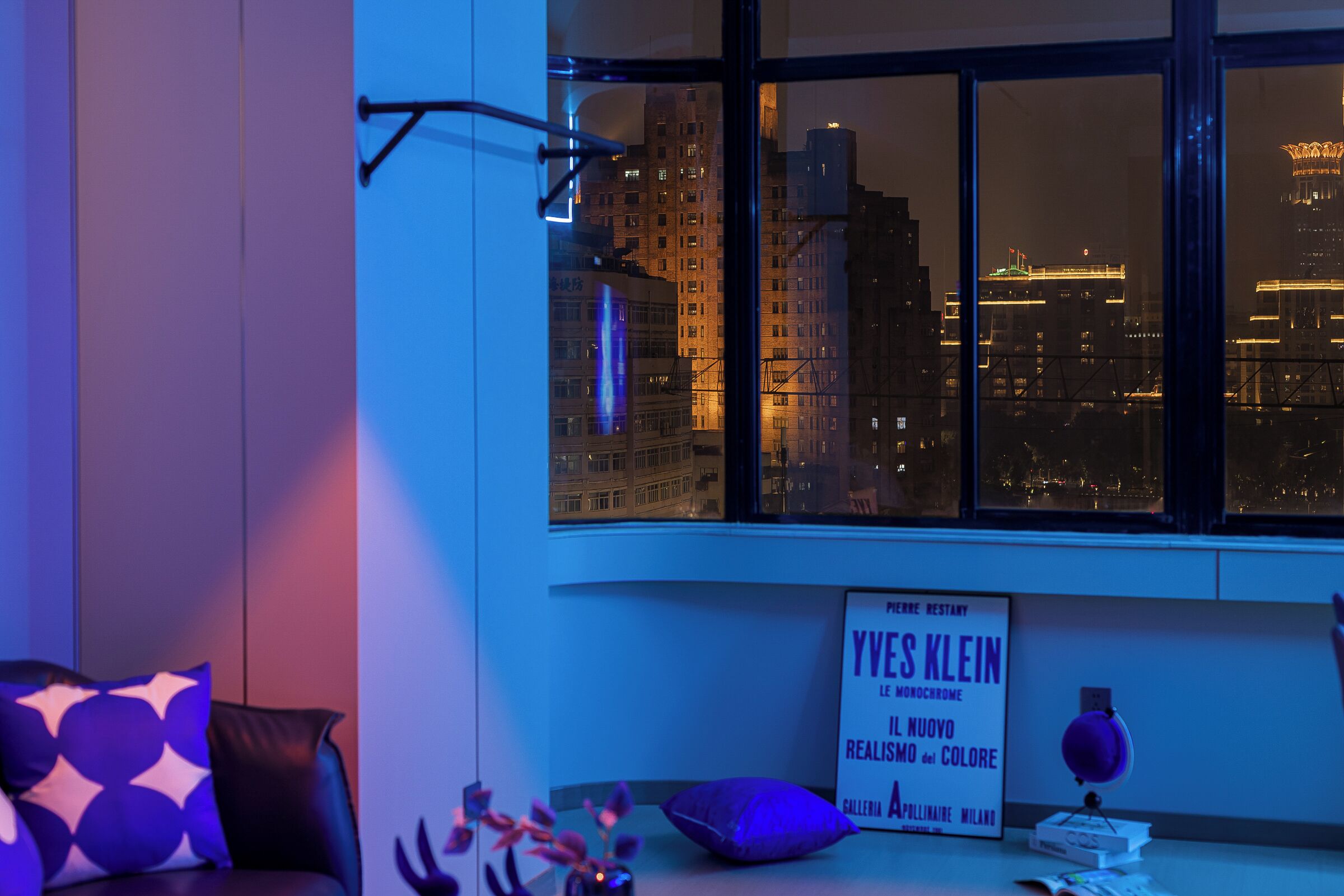 A room with a large window overlooking a cityscape at night, a sign for Yves Klein's gallery, and a lamp on the windowsill.