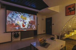A room with a large screen displaying a video game, a laptop, and a teddy bear on a table. - ShanghaiHills&SunflowerUltraHorizonHotel (Shanghai)