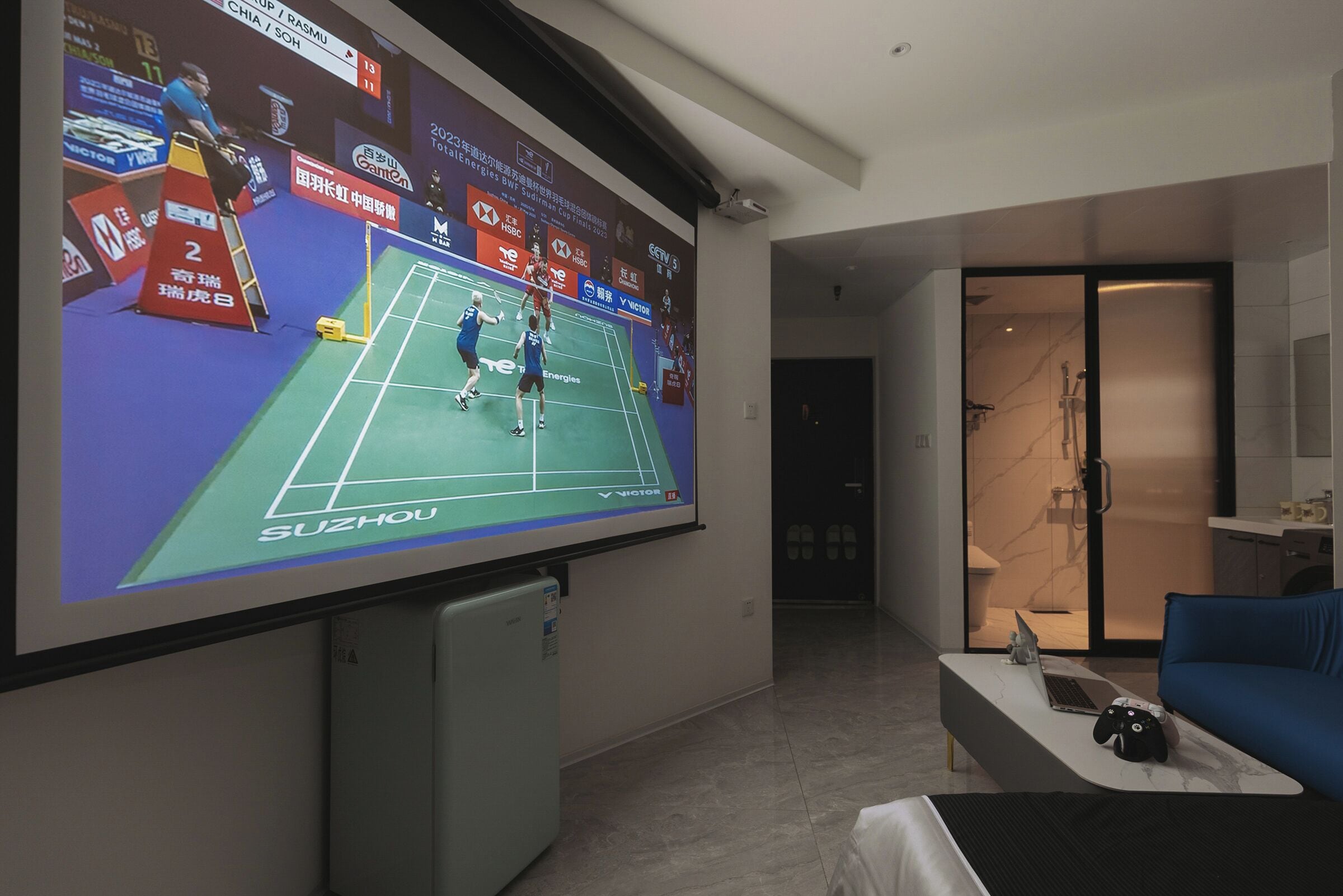 Habitación de hotel con una pantalla grande mostrando un partido de bádminton, una cama, un escritorio con una laptop y un control de videojuegos, y un área de ducha.
