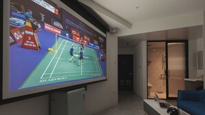 A hotel room with a large screen displaying a badminton match, a bed, a desk with a laptop and a game controller, and a shower area. - ShanghaiHills&SunflowerUltraHorizonHotel (Shanghai)