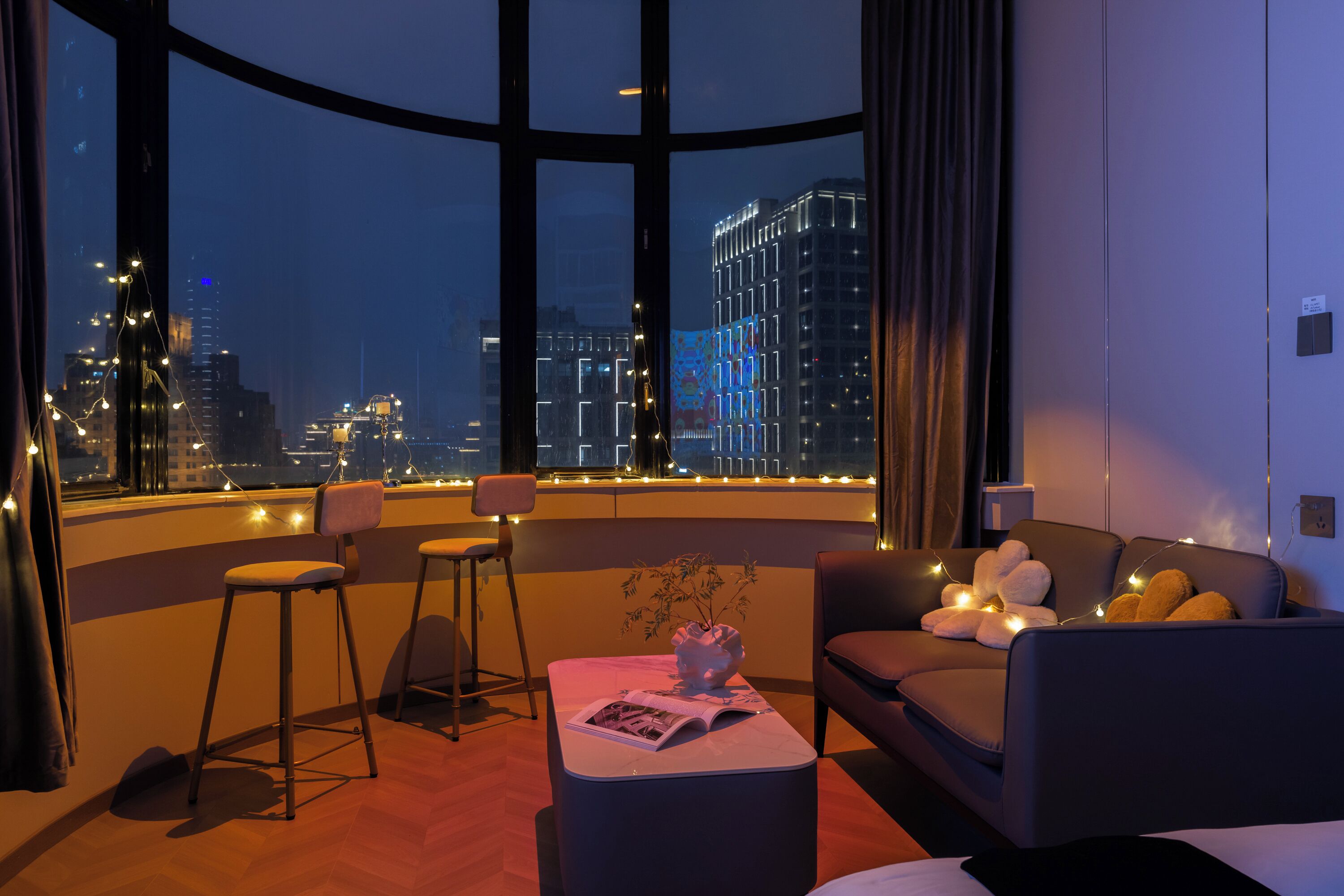 A modern hotel room with a curved window offering a city view, a sofa, a coffee table, and a bar stool.