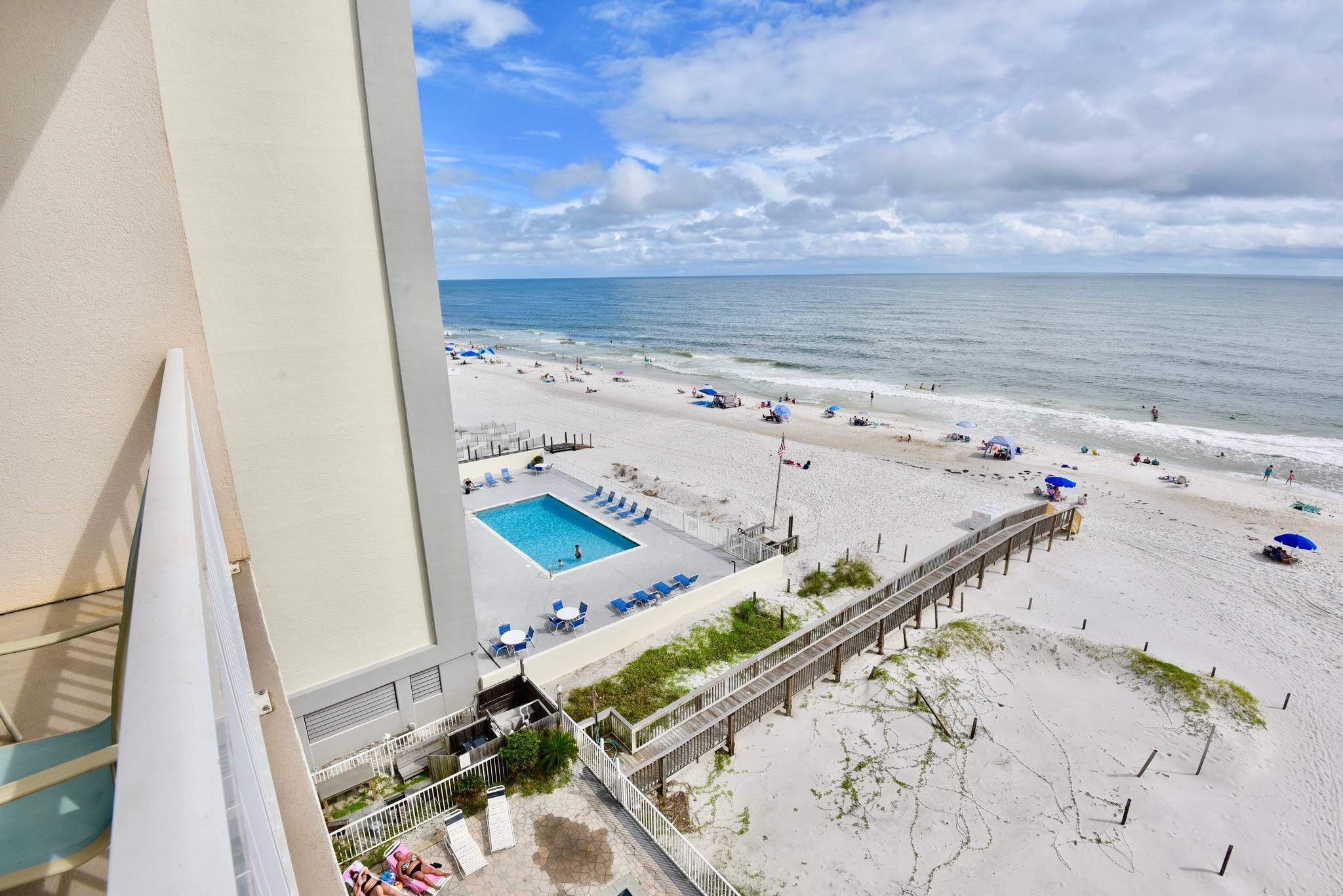 A beach view from a balcony with a pool, lounge chairs, and a wooden walkway.
