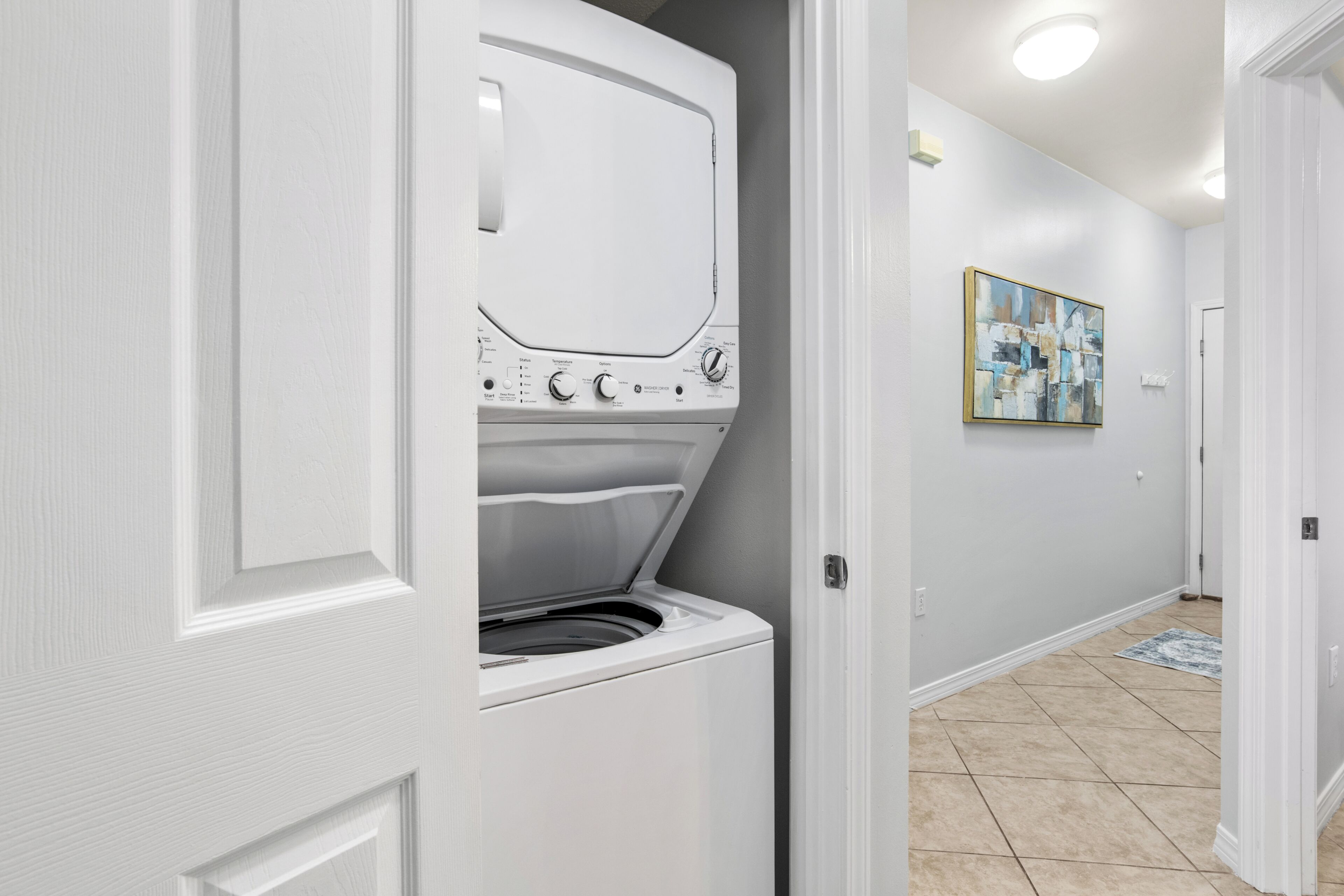 A laundry room with a front-loading washing machine and dryer, a door leading to another room, and a tiled floor.