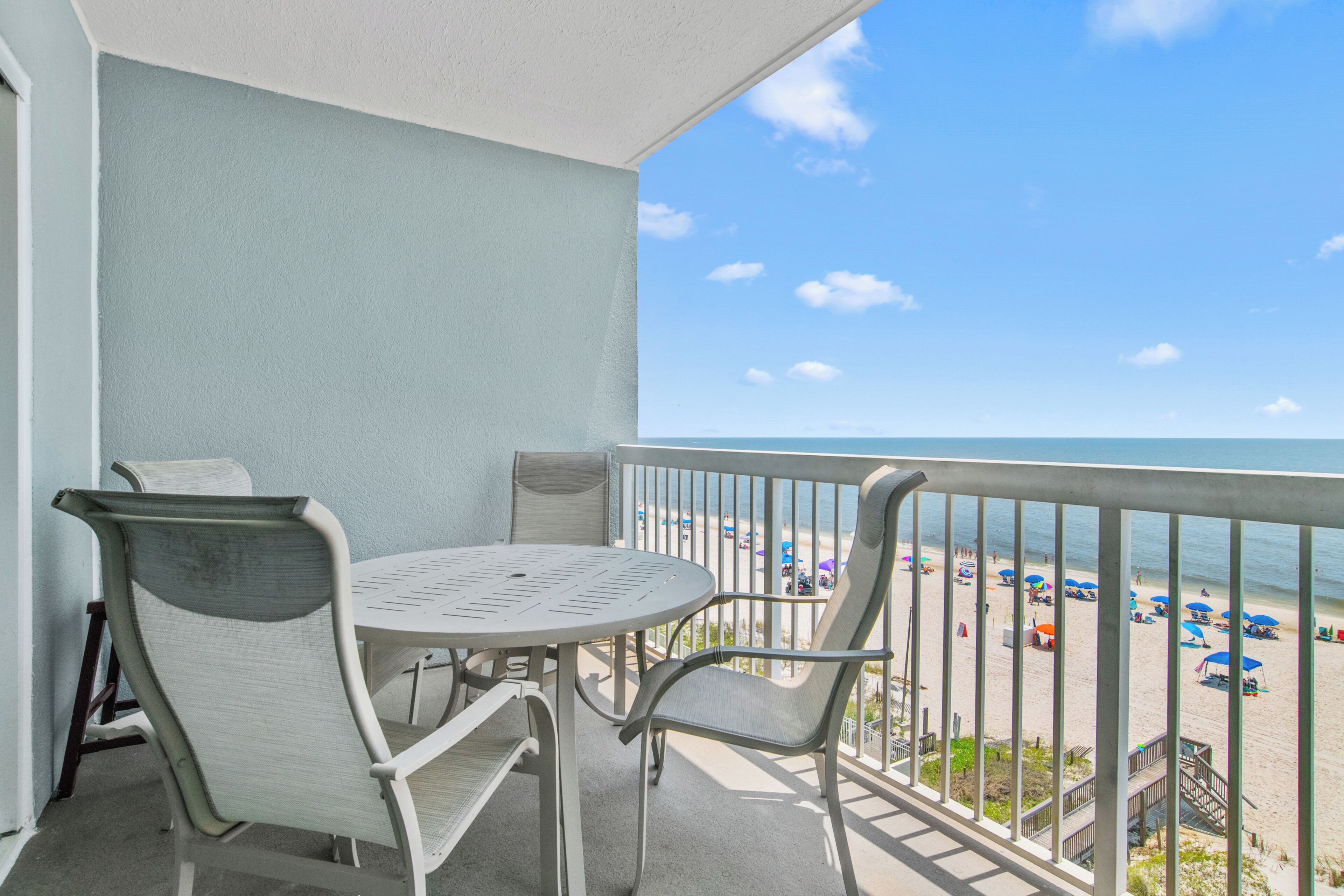 A balcony with a table and chairs, overlooking a beach with umbrellas and people.