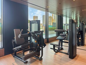 A modern gym with various exercise machines, large windows offering a view of the cityscape, and a clean, well-lit interior. - Bloomfields 1BR in Binghatti Onyx  (Dubai)