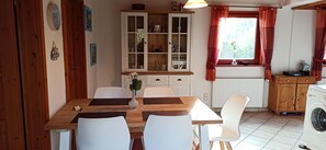 A dining area with a wooden table and white chairs, a cabinet with glass doors, and a window with curtains. - Vrbo Property (Windeby)