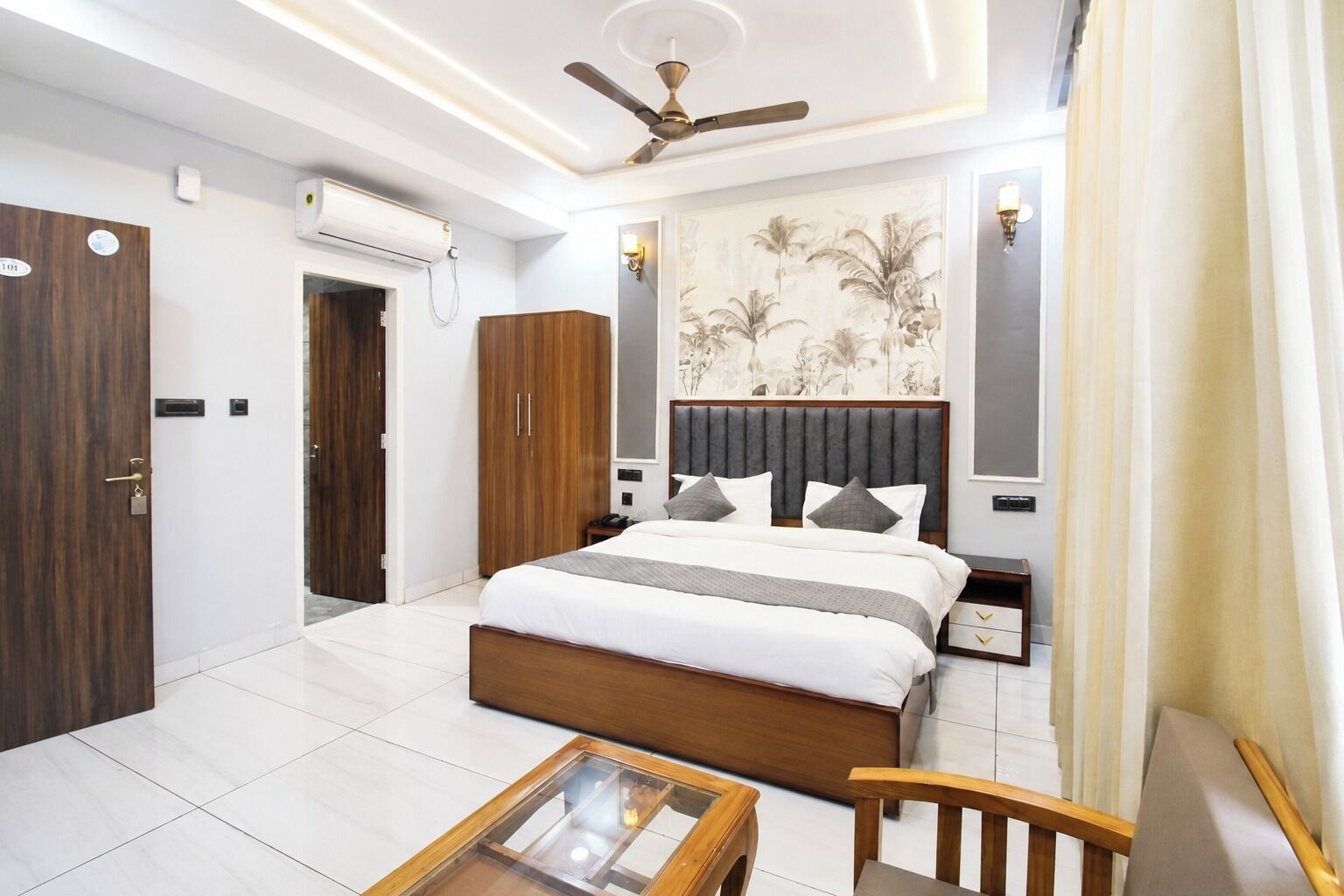 A hotel room with a bed, a desk, a chair, a ceiling fan, and a wall-mounted air conditioner.