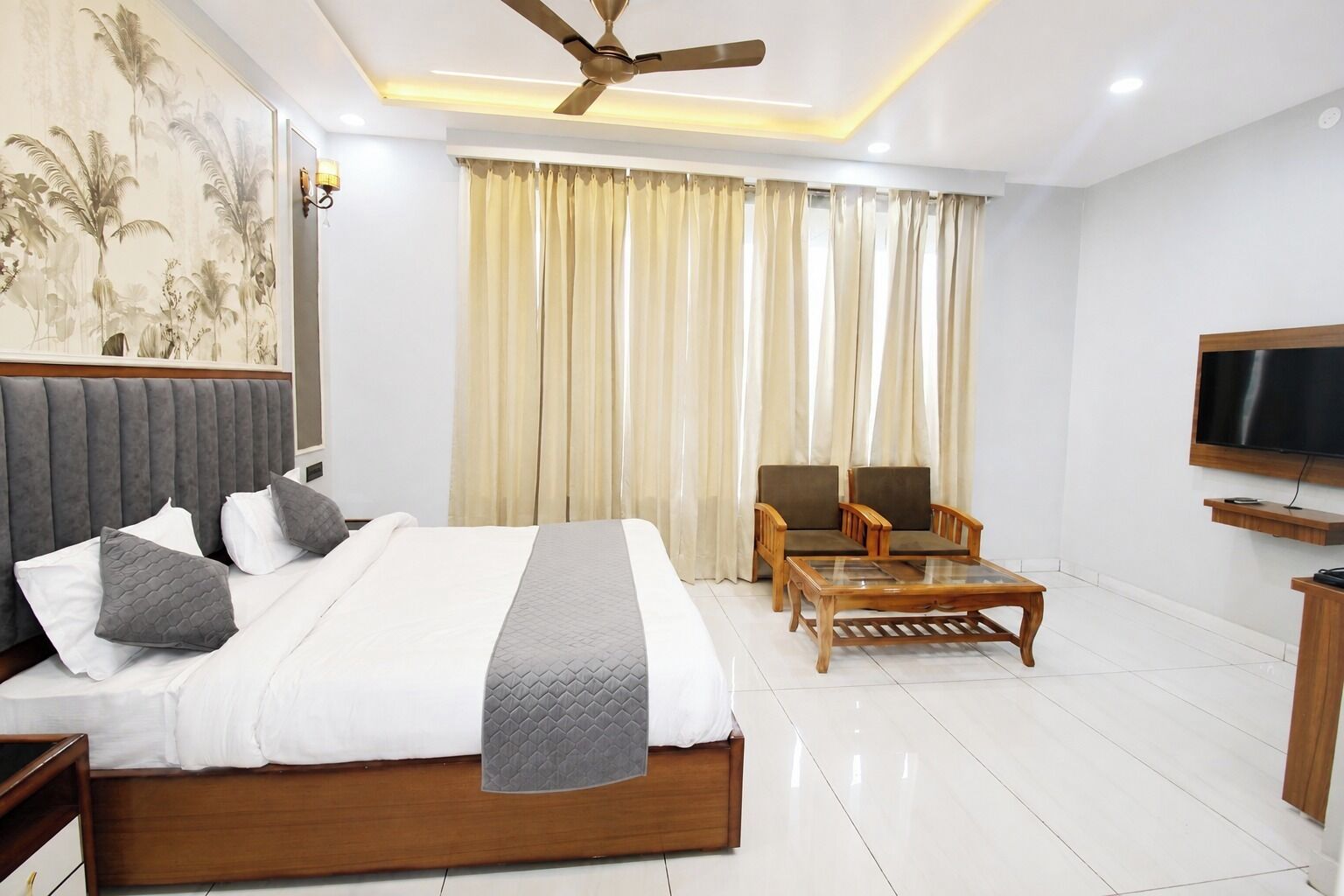 A hotel room with a large bed, two armchairs, a coffee table, a flat-screen TV, and a ceiling fan.