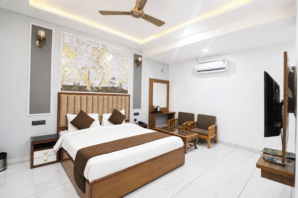 A hotel room with a large bed, a desk, two chairs, a ceiling fan, an air conditioning unit, and a television.