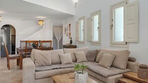 A living room with a beige sectional sofa, a wooden coffee table, and a dining area with a wooden table and chairs. - Solis Akrotiri Villa (Santorini)