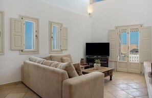 A living room with a sofa, a coffee table, a television, and a balcony door. - Solis Akrotiri Villa (Santorini)