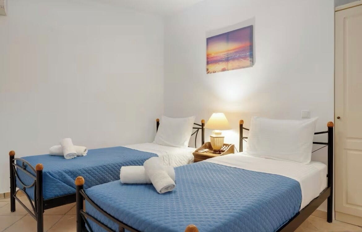 A hotel room with two beds, each with a blue bedspread and white pillows, a bedside table with a lamp, and a painting on the wall.