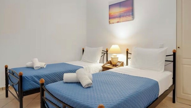 A hotel room with two beds, each with a blue bedspread and white pillows, a bedside table with a lamp, and a painting on the wall. - Solis Akrotiri Villa (Santorini)
