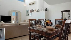 A dining area with a wooden table set for four, a sofa, and a television in the background. - Solis Akrotiri Villa (Santorini)