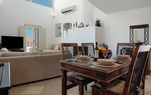 A dining area with a wooden table set for four, a sofa, and a television in the background. - Solis Akrotiri Villa (Santorini)