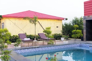 A pool area with lounge chairs, a small pool, and a building with a red roof. - Alaya Stays Royal Green Farm 2BHK W Pool (Gurugram)