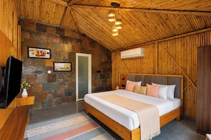 A bedroom with a stone wall, a bed with pillows, a television, and an air conditioning unit. - Alaya Stays Royal Green Farm 2BHK W Pool (Gurugram)