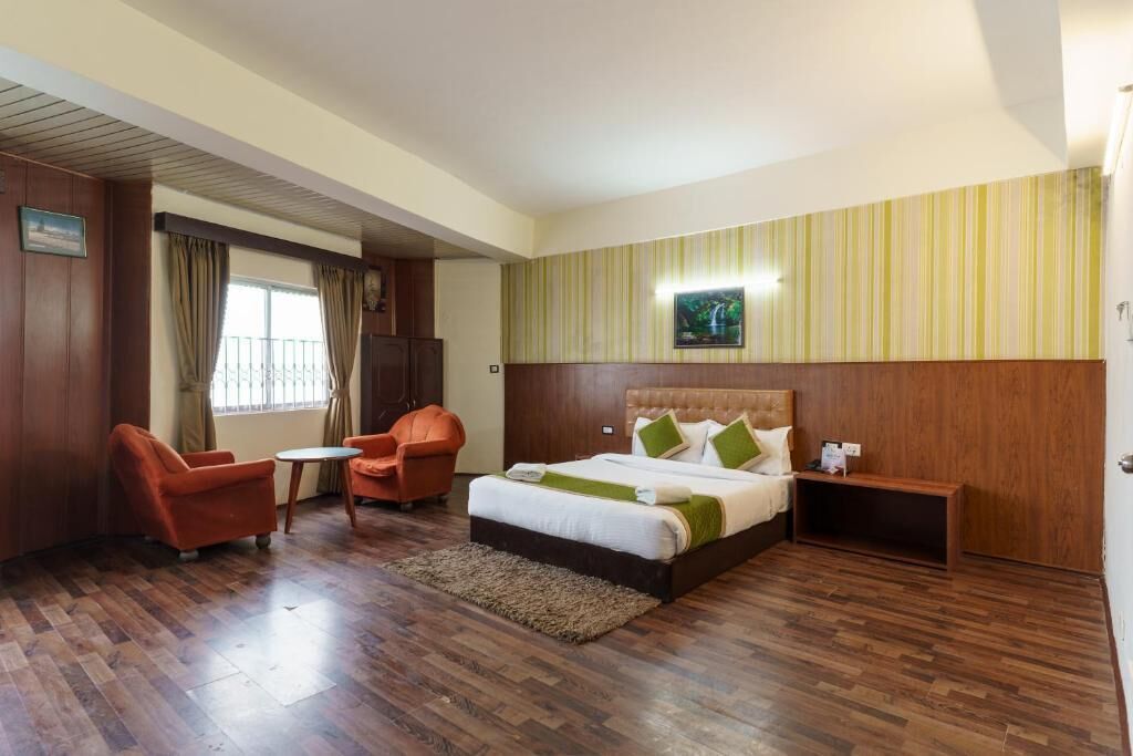 Une chambre d’hôtel comprenant un lit, deux fauteuils, une petite table, une table de chevet et une fenêtre avec des rideaux.