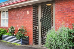 Ein rot backsteins Gebäude mit einer nummerierten Tür, einem Metallgitter und einem kleinen Garten mit Pflanzen.