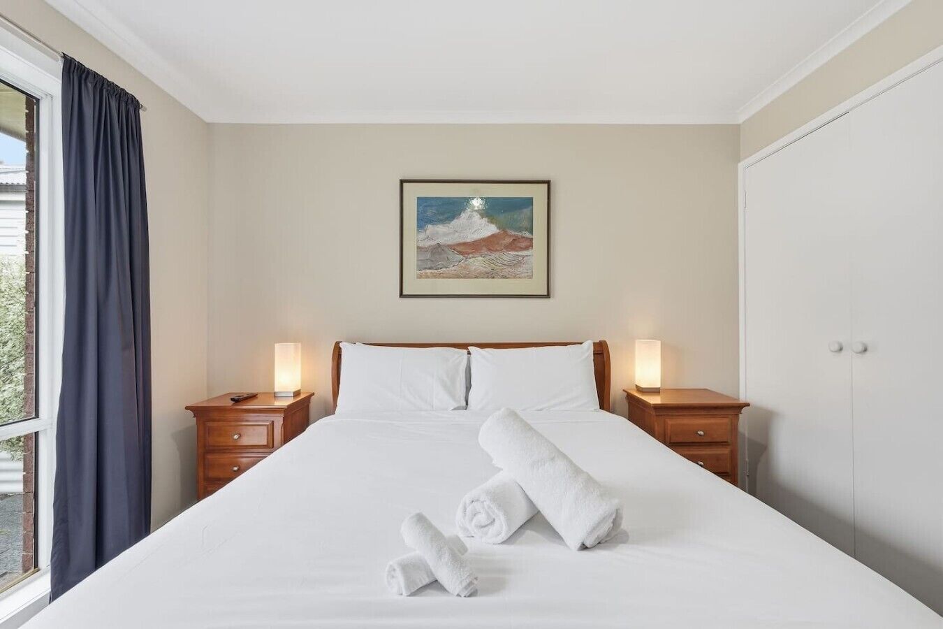 A hotel room with a large bed, two bedside tables with lamps, a window with curtains, and a framed picture on the wall.