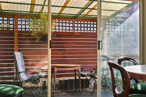 A covered outdoor seating area with a wooden table and chairs, a glass partition, and a view of a building through the glass. - King`s Corner Retreat (Sandy Bay)