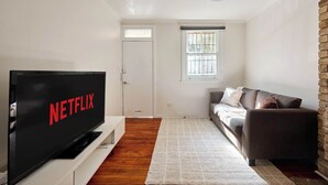 Ein Wohnzimmer mit Flachbildfernseher auf einem Schrank, einer Couch, einem Fenster mit Jalousien und einer Tür.
