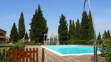 Una piscina con valla de madera, un pabellón y altos árboles al fondo.