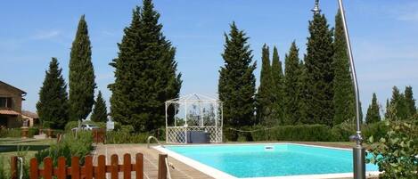 A swimming pool with a wooden fence, a gazebo, and tall trees in the background.