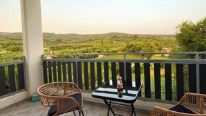 Ein Balkon mit Korbstühlen, einem kleinen Tisch und einer Laterne. Im Hintergrund sind grüne Hügel und ein entferntes Gebäude zu sehen.