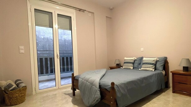 Ein Schlafzimmer mit einem Bett, einem Nachttisch, einer Lampe, einem Fenster mit Jalousien und einer Tür, die auf einen Balkon führt.