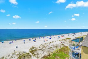 Ein Strand mit Sonnenschirmen und Liegestühlen, ein Gebäude mit einem Balkon und ein klarer Himmel.