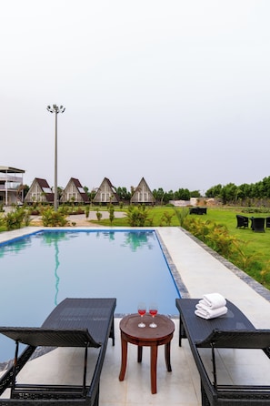 Uma piscina com espreguiçadeiras e uma mesa, rodeada por um jardim bem cuidado e uma fileira de edifícios tipo bungalow.