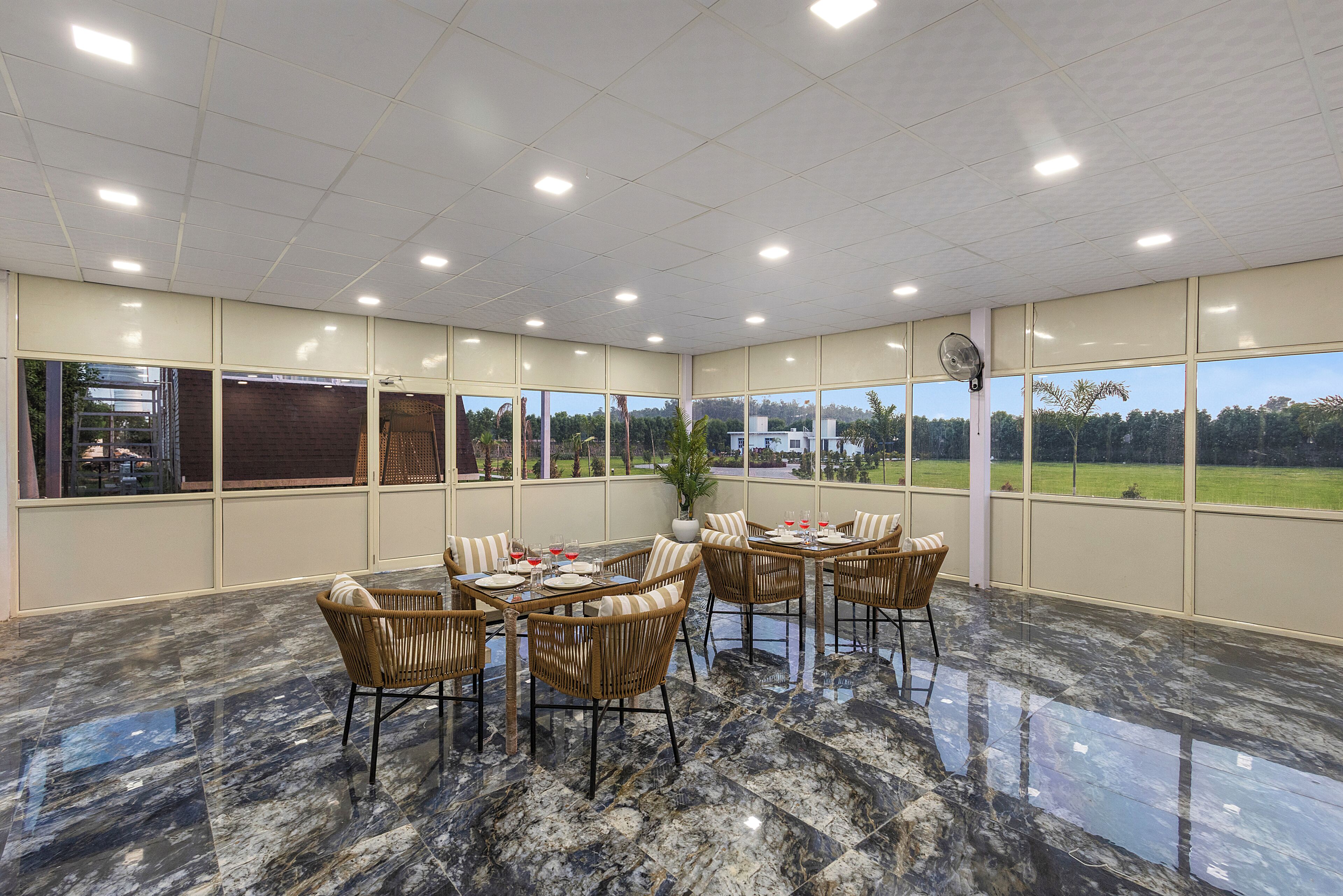 Un espace intérieur spacieux, avec une table à manger pour quatre personnes, entourée de chaises en osier, et de grandes fenêtres offrant une vue sur un paysage verdoyant.