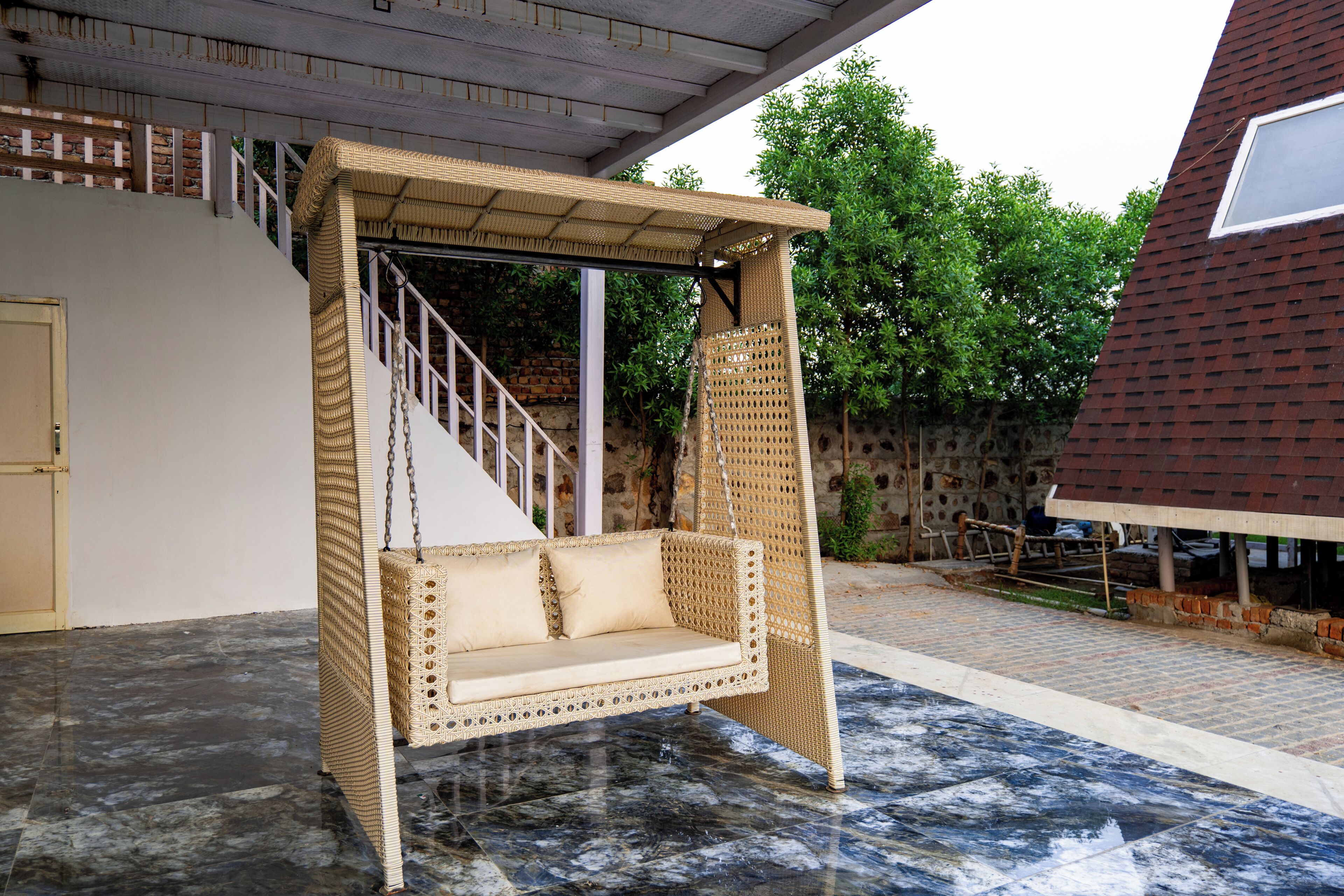 A wicker swing chair with a cushioned seat and backrest, suspended from a metal frame, set on a tiled patio.