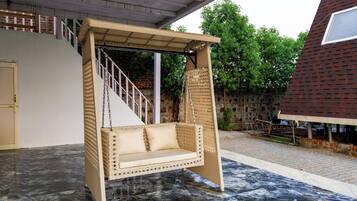 A wicker swing chair with a cushioned seat and backrest, suspended from a metal frame, set on a tiled patio.