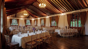 Un salón de banquetes con varias mesas redondas, piso de madera y grandes ventanales.